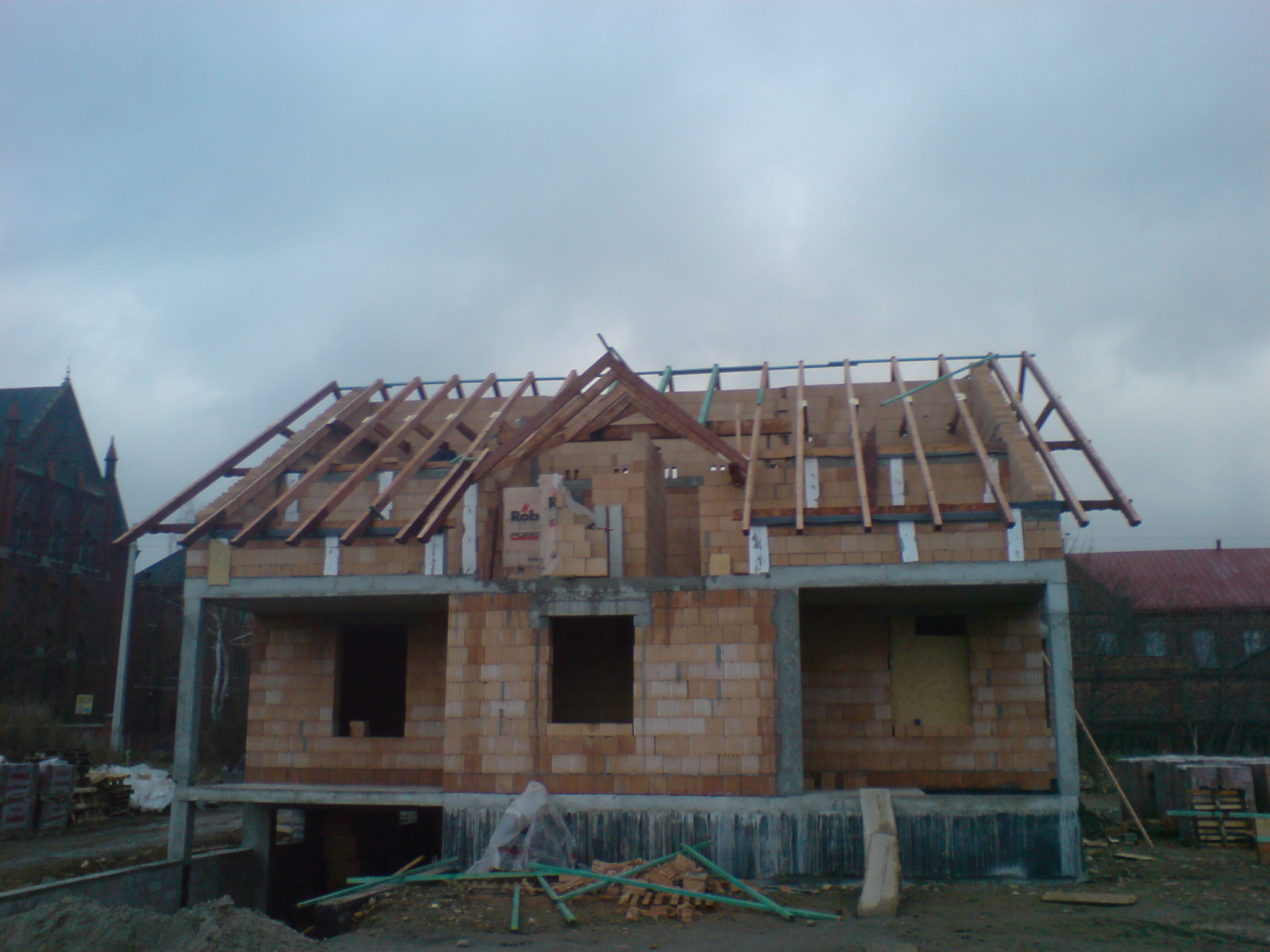 Budowa domu jednorodzinnego w stanie surowym otwartym: widoczne ściany z pustaków ceramicznych, więźba dachowa w trakcie montażu, zarys piwnicy i okoliczna zabudowa w tle.