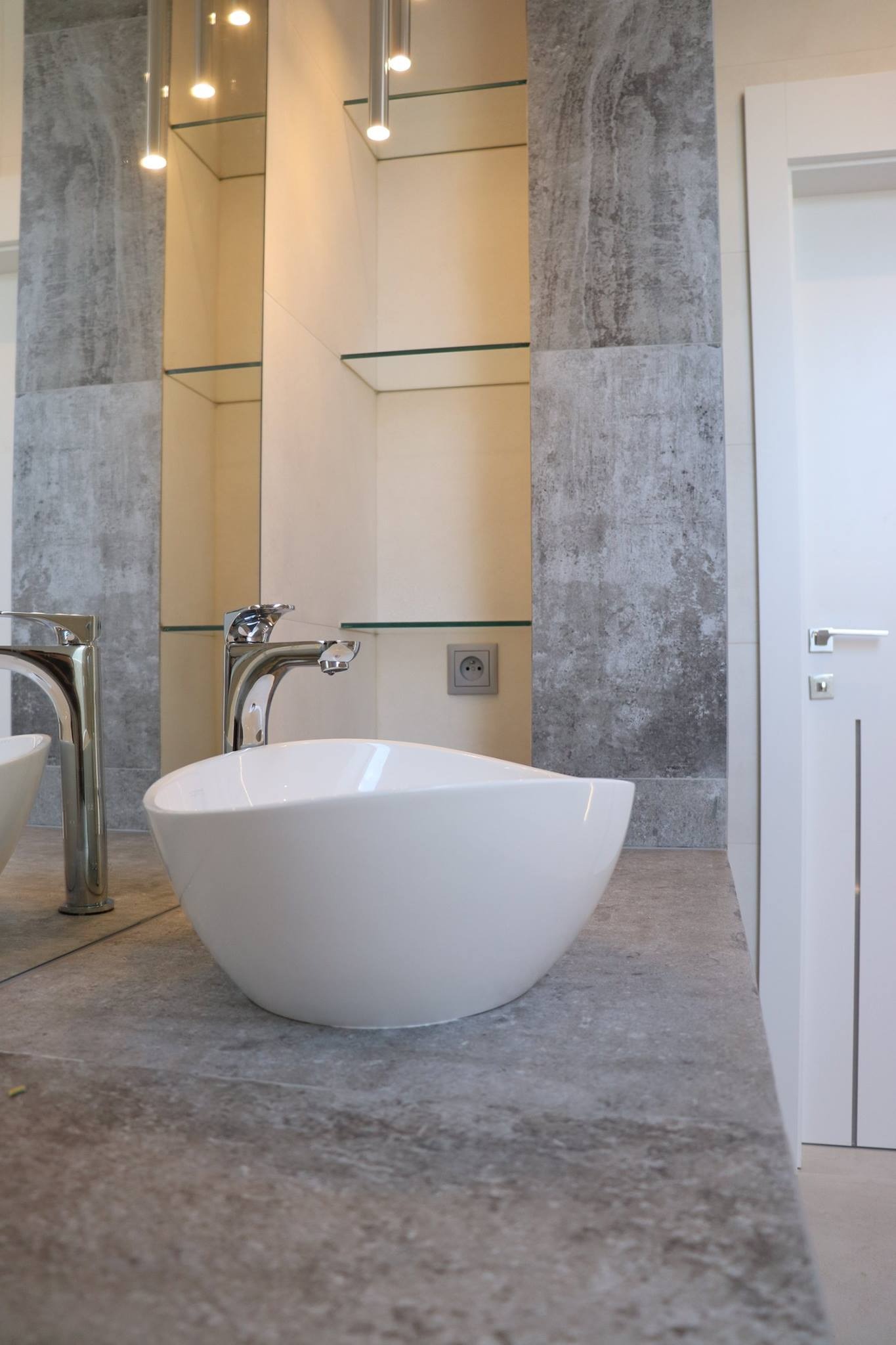 Nowoczesna łazienka z owalną białą umywalką nablatową, chromowaną baterią, lustrem, półkami i szarą okładziną ściany imitującą beton.