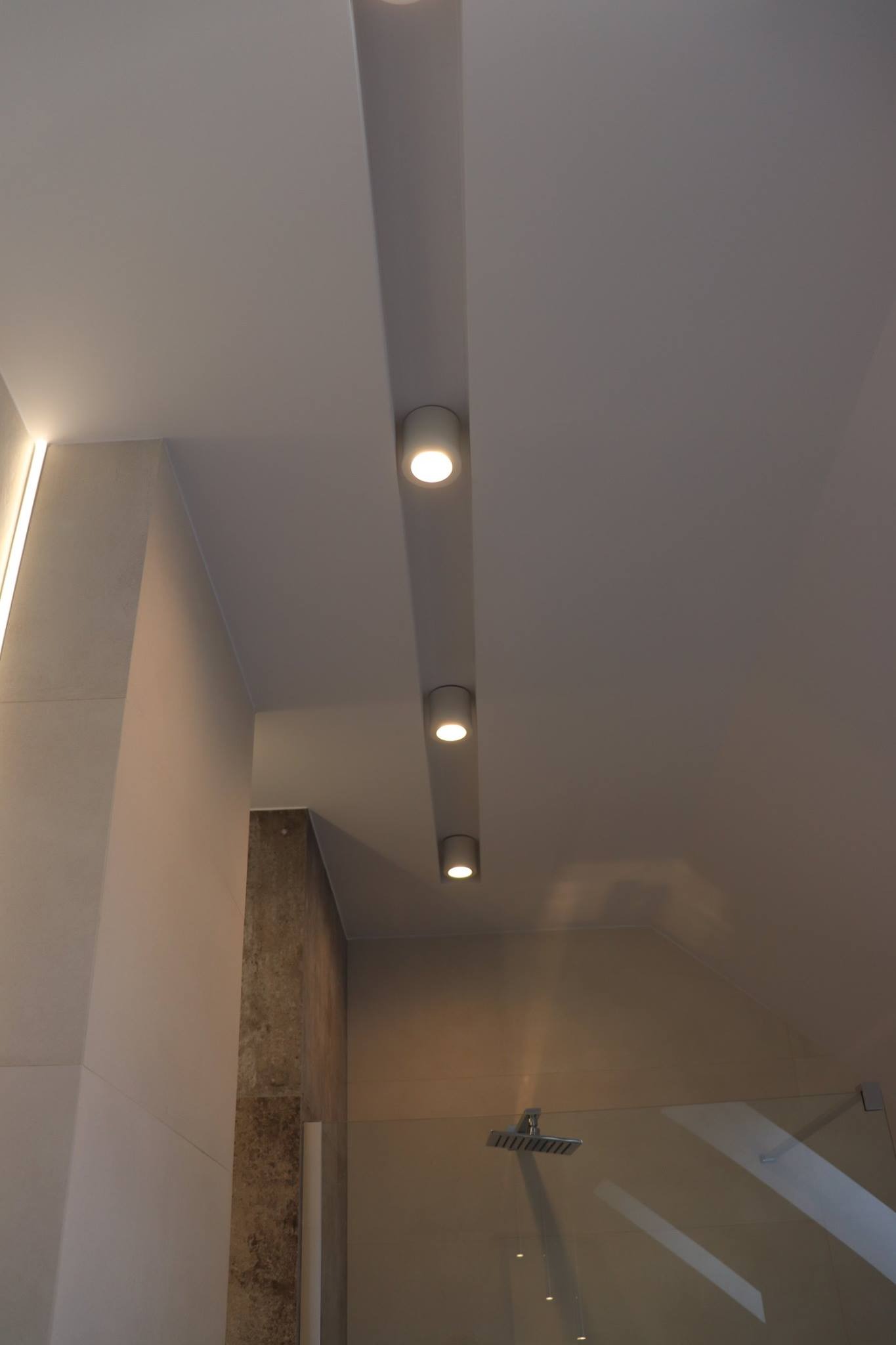 Nowoczesna łazienka z podwieszanym sufitem i oświetleniem punktowym LED, widoczna kabina prysznicowa z deszczownicą, jasne płytki na ścianach i dekoracyjny panel z kamienia.