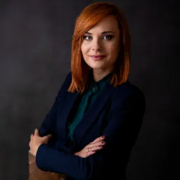 Portret kobiety w granatowym żakiecie i zielonej koszuli, z rudymi włosami, skrzyżowanymi rękami, opierającej się o drewniane krzesło na ciemnym tle.