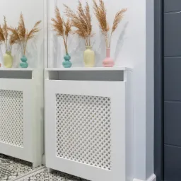 Biała, ażurowa obudowa grzejnika z półką, na której stoją trzy kolorowe wazoniki z suszonymi trawami, odbijająca się w lustrze; podłoga wyłożona wzorzystymi płytkami.