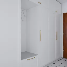 Biała zabudowa przedpokoju z szafami i siedziskiem, złote uchwyty i wieszak na ubrania, podłoga w biało-czarną szachownicę.