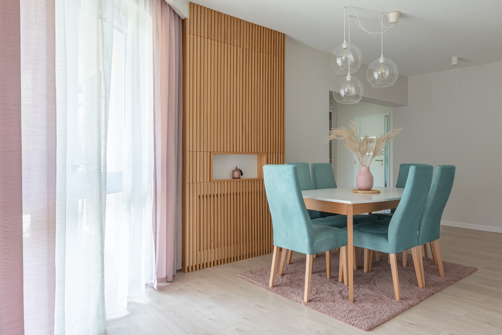 Jasne wnętrze jadalni z drewnianą ścianą z lamelami, wnęką dekoracyjną i turkusowymi krzesłami wokół białego stołu, oświetlone wiszącymi lampami z przezroczystego szkła.