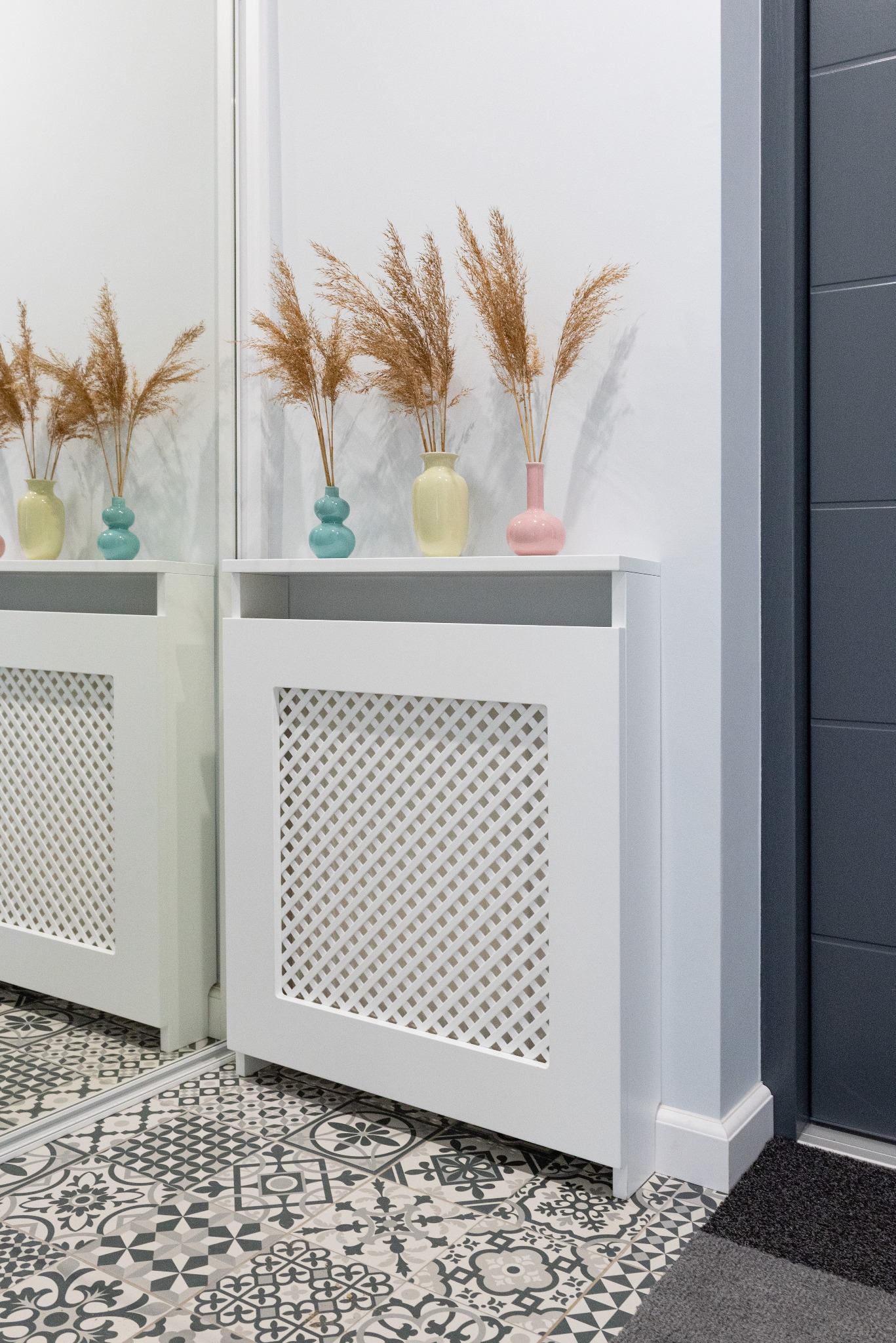 Biała, ażurowa obudowa grzejnika z półką, na której stoją trzy kolorowe wazoniki z suszonymi trawami, odbijająca się w lustrze; podłoga wyłożona wzorzystymi płytkami.