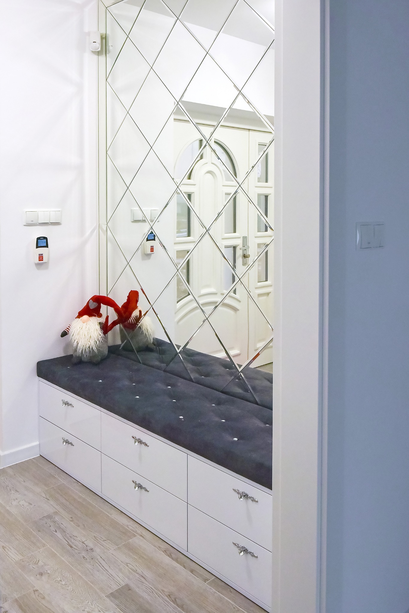 Biała szafka z szufladami i szarym siedziskiem, udekorowana pluszowymi skrzatami, na tle ściany z lustrami w romby, odbijającymi białe drzwi wejściowe.