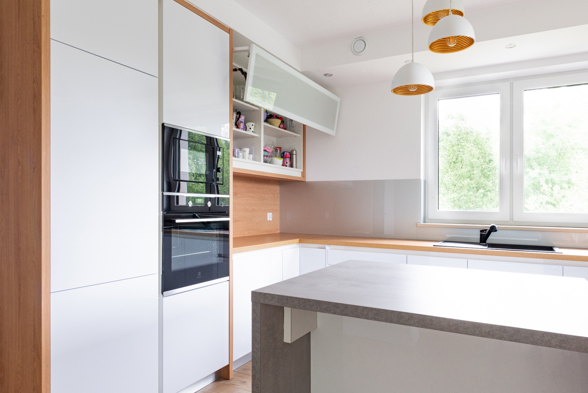 Nowoczesna, jasna kuchnia z białą zabudową, drewnianymi akcentami i wyspą z szarym blatem. Widoczny otwarty górny szafka z półkami, piekarnik w zabudowie i okno z widokiem na zieleń.