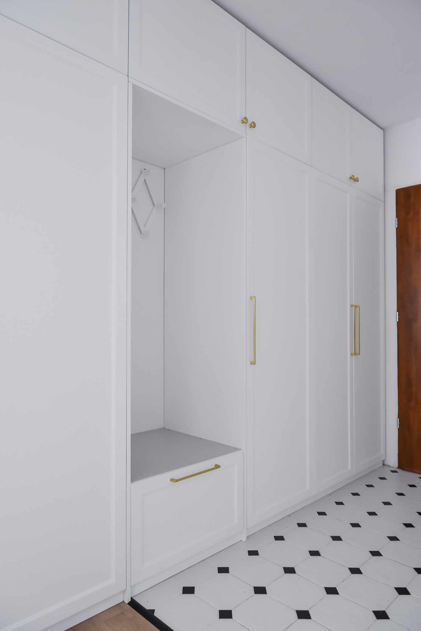 Biała zabudowa przedpokoju z szafami i siedziskiem, złote uchwyty i wieszak na ubrania, podłoga w biało-czarną szachownicę.
