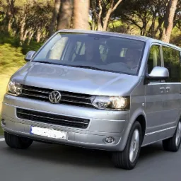 Srebrny Volkswagen Transporter T5 na drodze w otoczeniu drzew, z widocznymi dwoma osobami wewnątrz.
