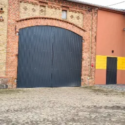 Duża, metalowa brama garażowa w łukowym przejściu ceglanego budynku, obok fragment budynku z pomarańczową elewacją i żółtym pasem, na brukowanym podwórzu.