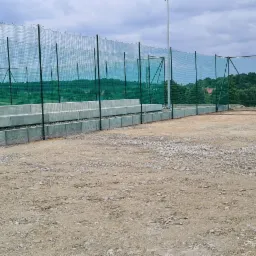 Nowo wybudowane ogrodzenie panelowe z siatki w kolorze zielonym, zabezpieczające teren sportowy z widocznymi betonowymi elementami konstrukcyjnymi i podłożem z gruzu.