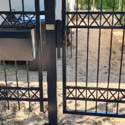 Częściowo otwarta, czarna brama wjazdowa z metalowymi prętami i ozdobnym wzorem krzyżyków w górnej części, z zamontowaną skrzynką na listy, widok na podwórko w tle.