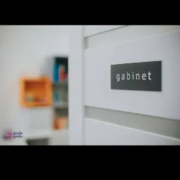 Białe drzwi z szarą tabliczką z napisem 'gabinet' i rozmazane wnętrze z kolorowymi półkami w tle.