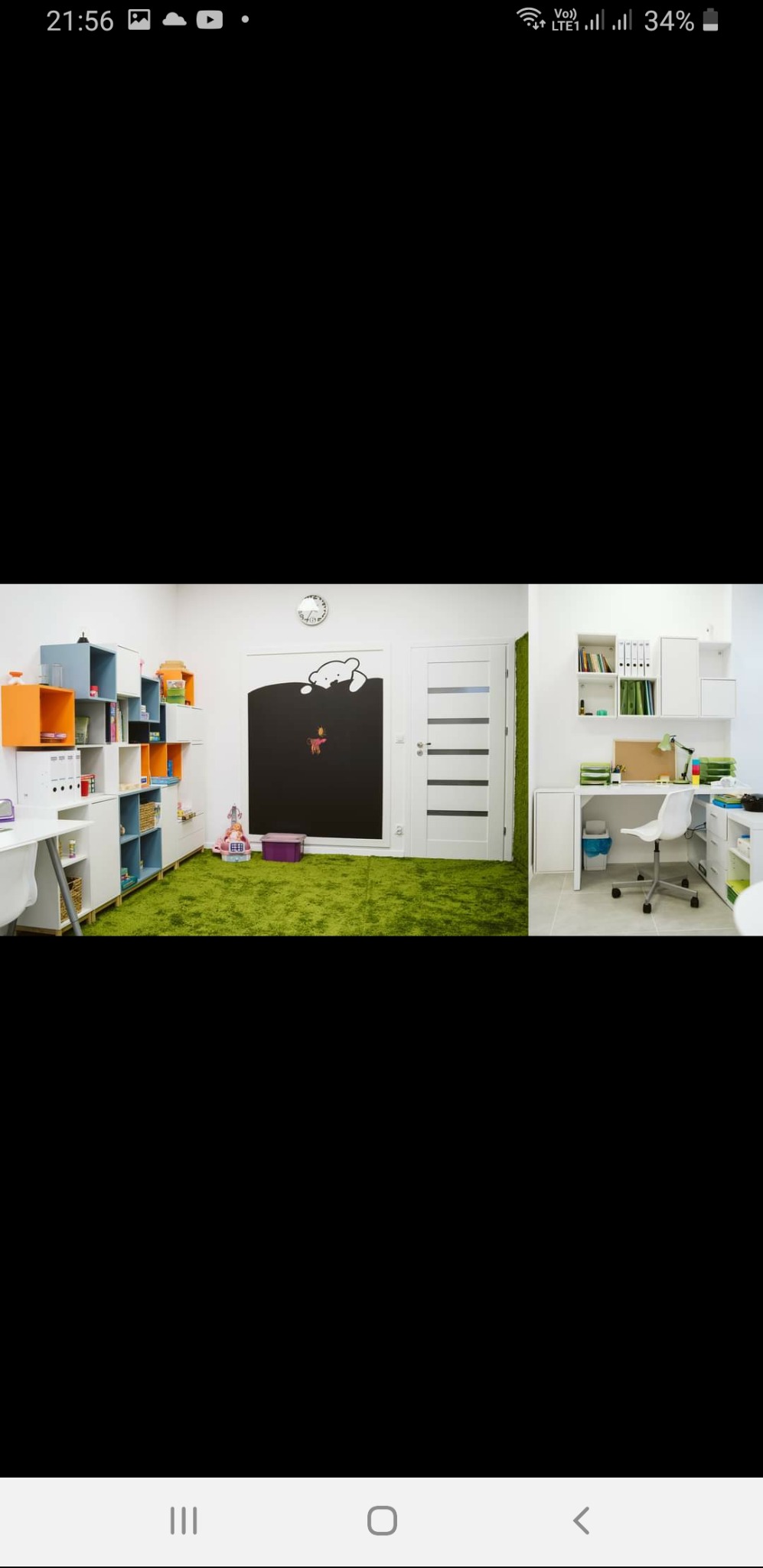 Jasny pokój terapeutyczny z zielonym dywanem imitującym trawę, czarną tablicą z rysunkami i meblami do przechowywania z kolorowymi akcentami.