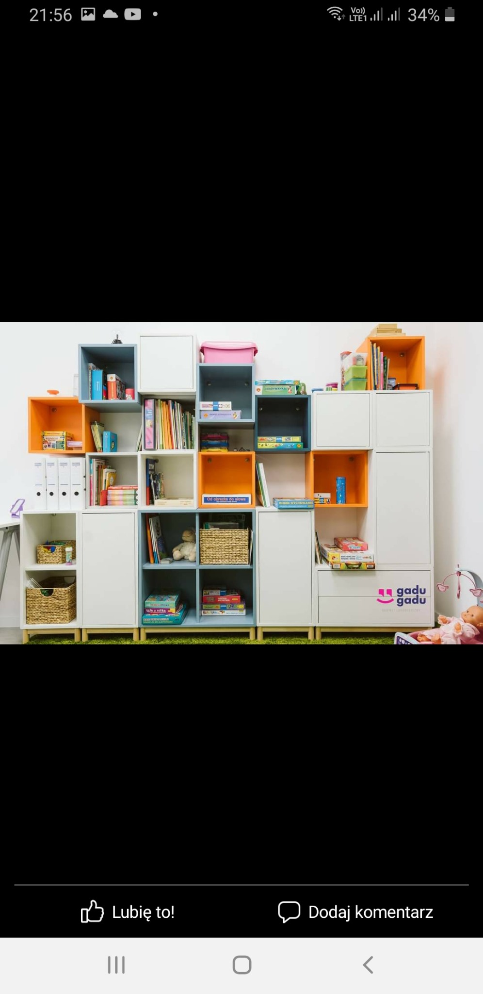 Modułowy regał dziecięcy z kolorowymi półkami, wypełniony książkami, grami i zabawkami, na zielonym dywanie, obok leży lalka.