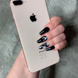 Dłoń z czarnymi, długimi paznokciami z zimowymi zdobieniami (śnieżynki, renifer) trzymająca telefon iPhone w kolorze różowego złota na tle szarego, futrzanego koca.