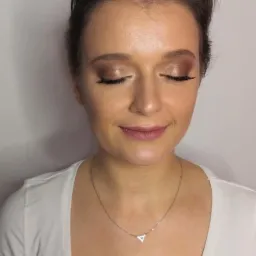 Portret kobiety z delikatnym, brązowym makijażem oczu i ust, z zamkniętymi oczami i subtelnym uśmiechem, noszącej białą koszulkę i srebrny naszyjnik z trójkątną zawieszką.