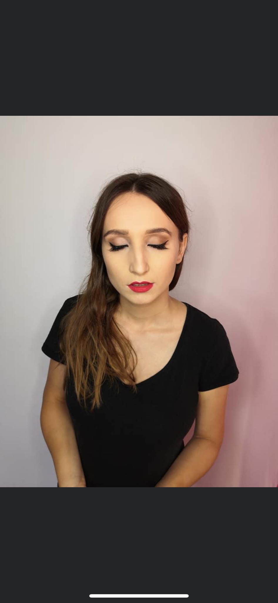 Portret kobiety z delikatnym makijażem oczu w stylu cat-eye i intensywnie czerwoną szminką na ustach, włosy częściowo opadające na czarną bluzkę z dekoltem w serek, na tle jasnej ściany z lekkim...