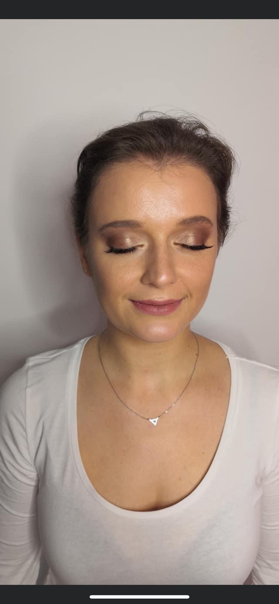 Portret kobiety z delikatnym, brązowym makijażem oczu i ust, z zamkniętymi oczami i subtelnym uśmiechem, noszącej białą koszulkę i srebrny naszyjnik z trójkątną zawieszką.