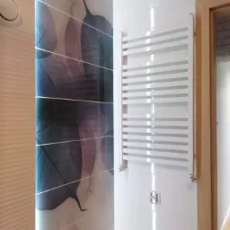 Biały grzejnik drabinkowy zamontowany na białej ścianie w łazience z dekoracyjnymi płytkami z motywem liści, widoczne gniazdko elektryczne i fragment drewnianej ościeżnicy.