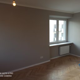 TAK-RÓB Jakub Żelazowski - Pokój po malowaniu ścian na szaro, widoczna podłoga z parkietu w jodełkę, okno z białymi ramami i grzejnik, w rogu niebieski worek z akcesoriami malarskimi.