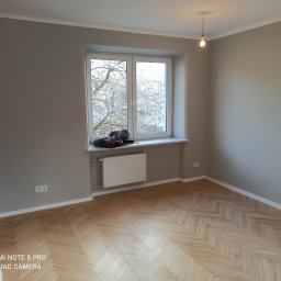 TAK-RÓB Jakub Żelazowski - Pokój po malowaniu ścian na szaro, z parkietem w jodełkę, białym oknem, grzejnikiem i pojedynczą żarówką na kablu.