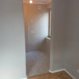 Widok przez drzwi do pomieszczenia z szarymi ścianami, terazzo na podłodze i pojedynczą żar&oacute;wką