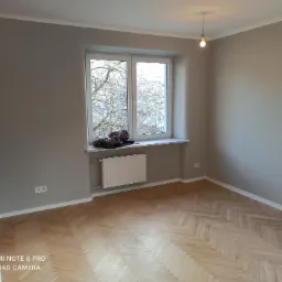 Pomalowane na szaro wnętrze pokoju z parkietem w jodełkę, białym oknem z widokiem na drzewa i wiszącą żarówką bez klosza.