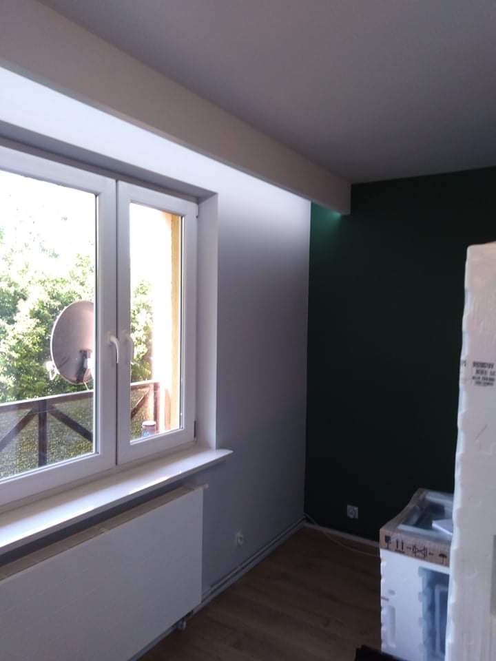 Pokój po malowaniu: ciemnozielona ściana, okno z widokiem na balkon z anteną satelitarną, grzejnik pod oknem, drewniana podłoga.