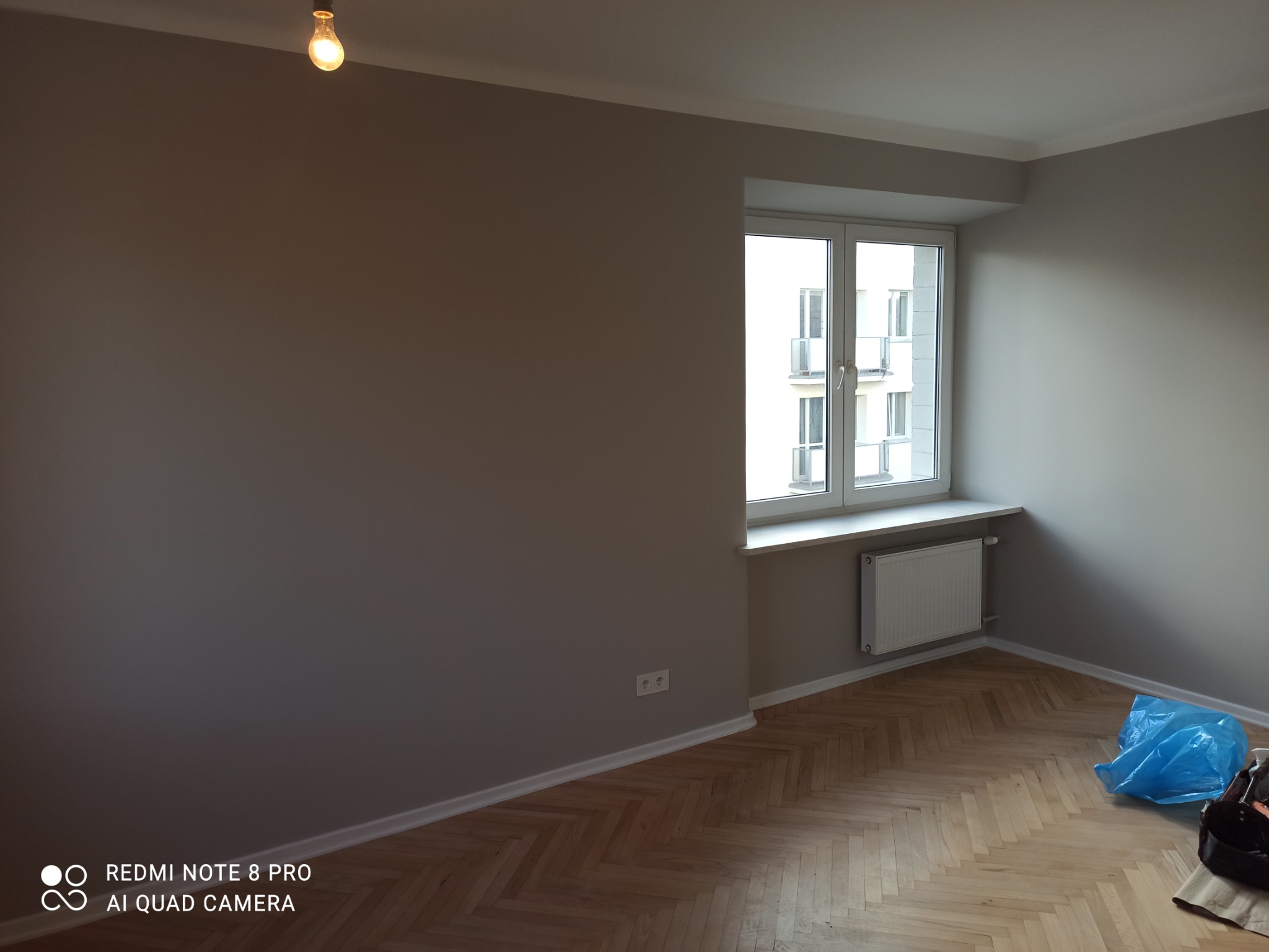 Pokój po malowaniu ścian na szaro, widoczna podłoga z parkietu w jodełkę, okno z białymi ramami i grzejnik, w rogu niebieski worek z akcesoriami malarskimi.