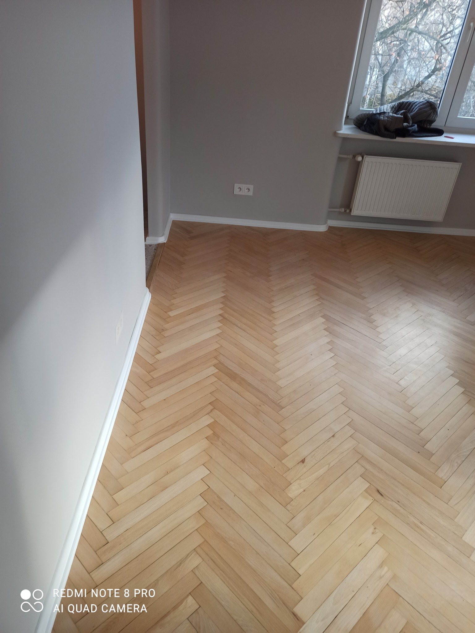 Parkiet ułożony w jodełkę w odnowionym pokoju, szare ściany, białe listwy przypodłogowe, grzejnik pod oknem