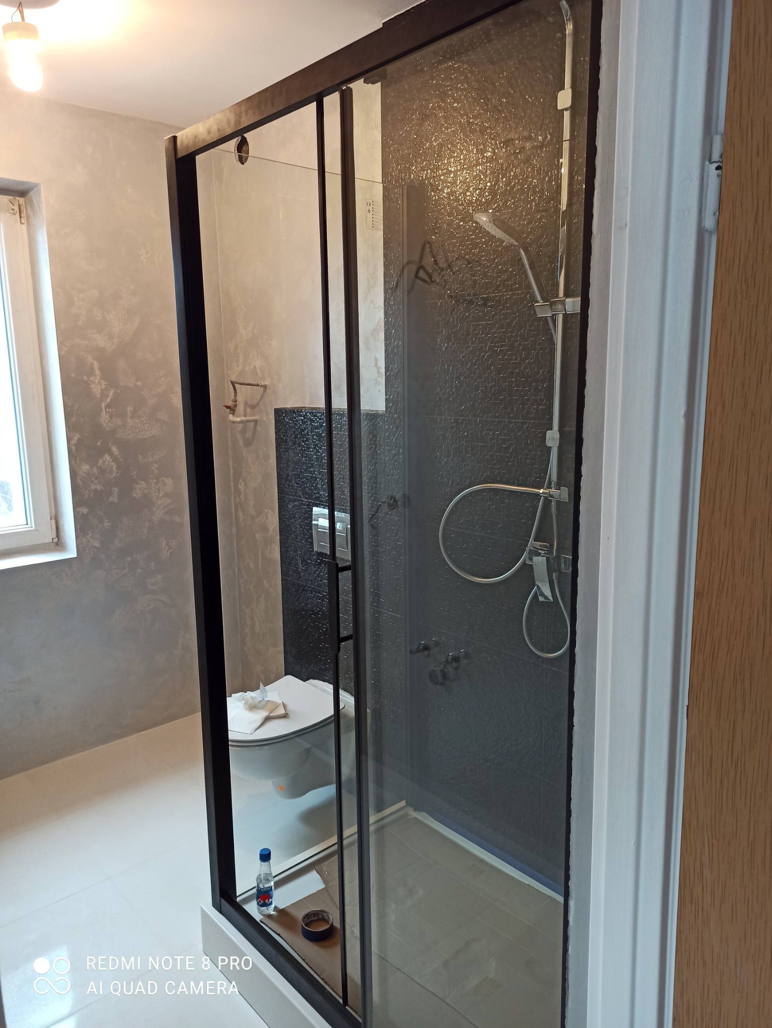 Nowoczesna kabina prysznicowa z czarnymi profilami i deszczownicą, widok z zewnątrz na toaletę i szpachlowane ściany w łazience.