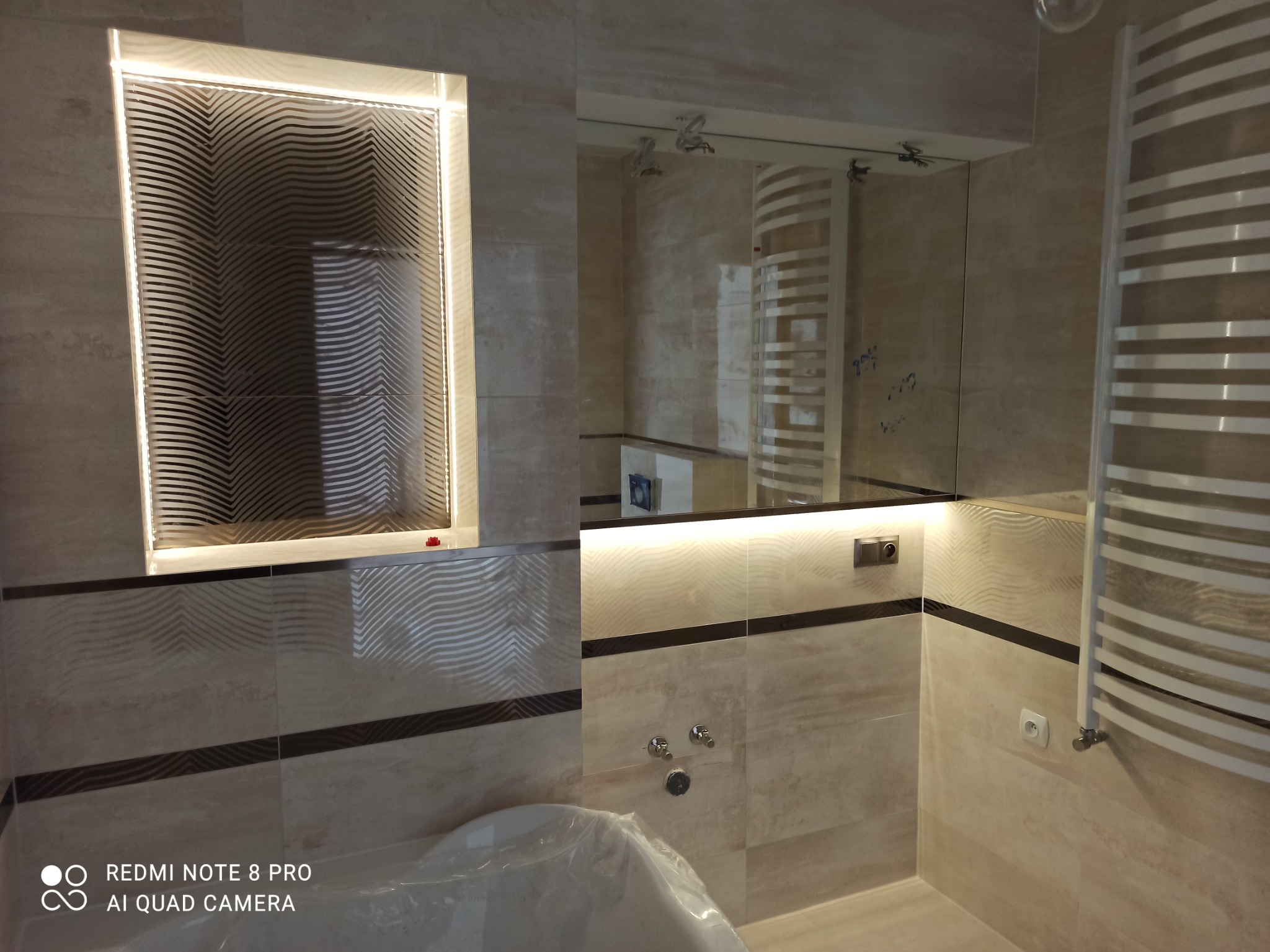 Wykończona łazienka w Jakubowie z podświetlanym lustrem o geometrycznym wzorze i grzejnikiem drabinkowym, widoczne płytki w odcieniach beżu z ciemnym pasem.