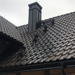 Nowy dach z ciemnej dachówki ceramicznej, z zamontowanymi stopniami kominiarskimi i nowoczesnym, kwadratowym kominem w kolorze czarnym, widoczne rynny.