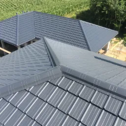 Nowoczesny, grafitowy dach z blachy trapezowej, widok z góry, z fragmentem komina i budynkiem w stanie surowym otwartym w tle, otoczonym zielenią.