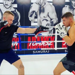 Dwóch mężczyzn w ringu bokserskim podczas sparingu, jeden z ochraniaczami na rękach, drugi w rękawicach bokserskich, w tle plakaty z wizerunkami bokserów.