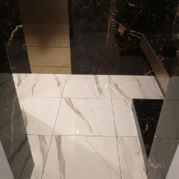 Wykończona łazienka z kontrastującym połączeniem białych płytek podłogowych imitujących marmur z ciemnymi, marmurkowymi płytkami na ścianach oraz elementami w kolorze taupe.