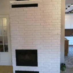 Biały, ceglany kominek z czarnym wkładem, widoczny fragment wnętrza z drewnianymi belkami stropowymi i białą zabudową kuchenną w tle.