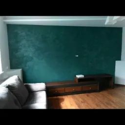 Ściana w salonie pomalowana na ciemny, morski odcień z efektem dekoracyjnym, widoczny szary narożnik i drewniana szafka RTV na parkiecie.