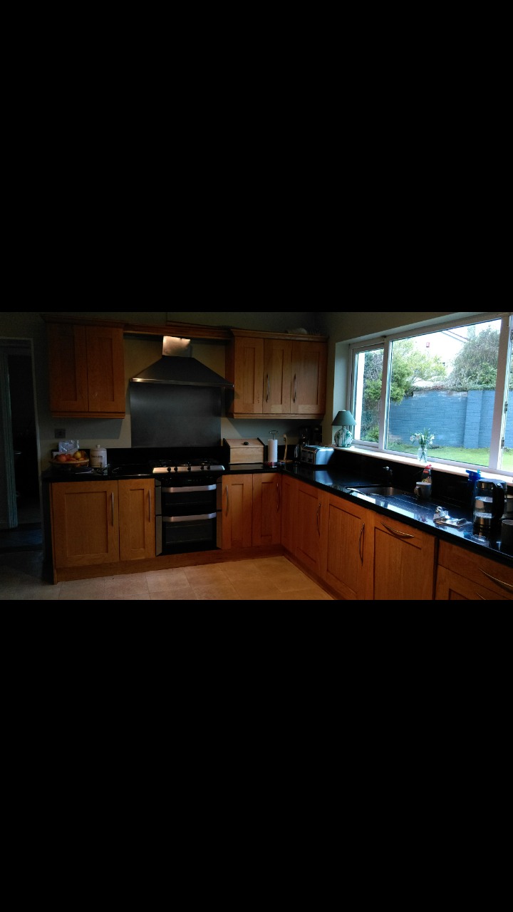 Kuchnia z drewnianymi szafkami, czarnym blatem i płytkami podłogowymi w odcieniach beżu, widok na ogród przez okno.