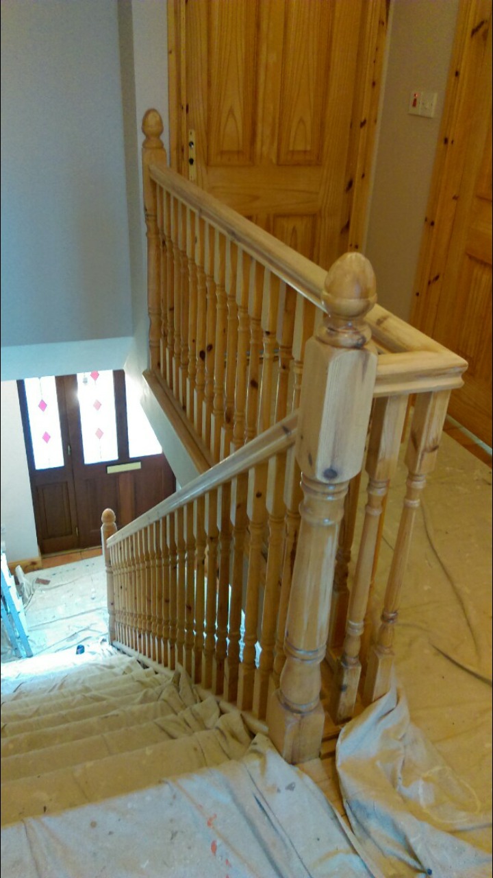Drewniana balustrada schodów zabezpieczona folią malarską podczas prac remontowych, widok z góry na klatkę schodową z drewnianymi drzwiami wejściowymi.