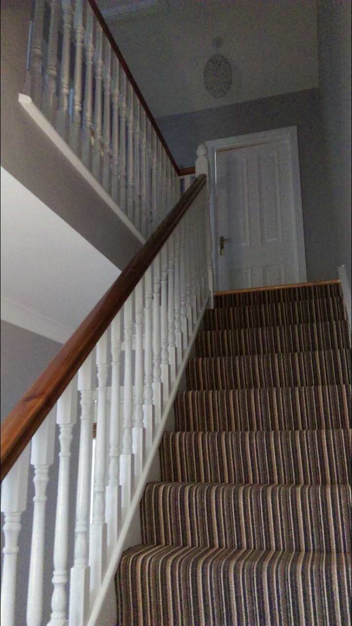 Schody z brązowo-beżową wykładziną w pionowe pasy, biała balustrada z drewnianym poręczą, białe drzwi na końcu biegu schodów, szara ściana.