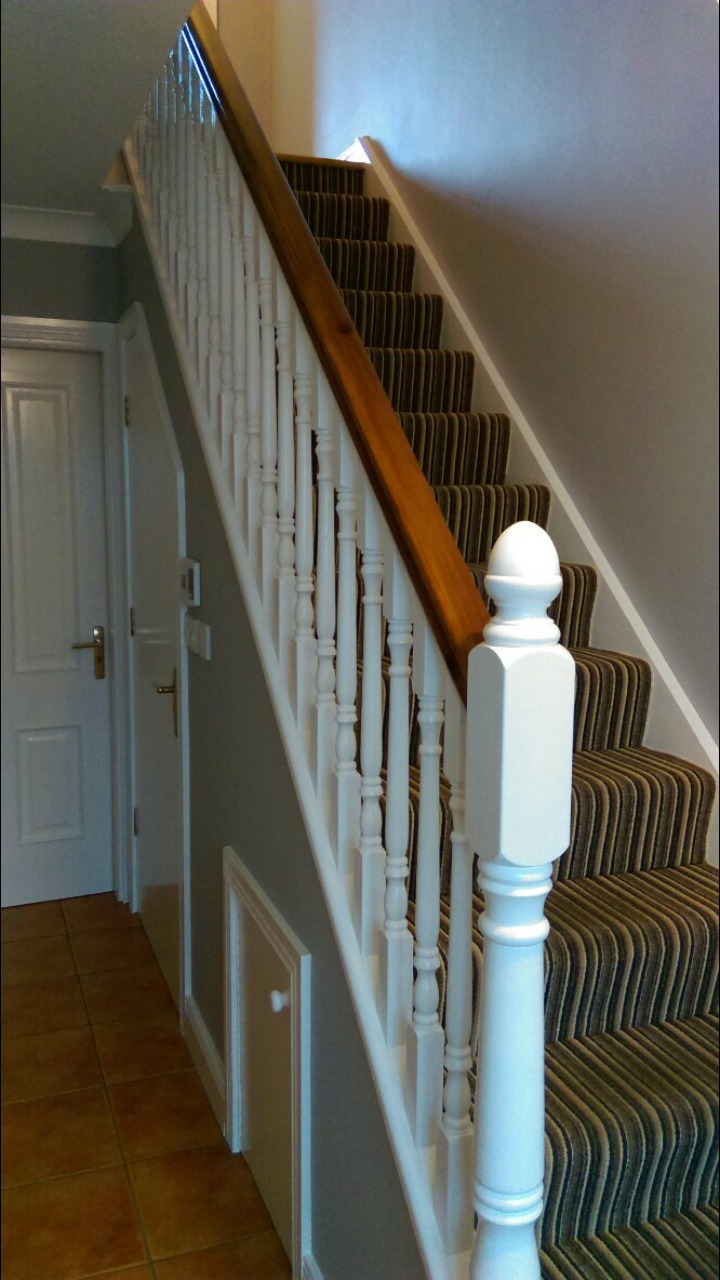 Klatka schodowa z drewnianą poręczą i białymi balustradami, wyłożona dywanem w brązowe pasy, widok z dołu na pierwsze stopnie