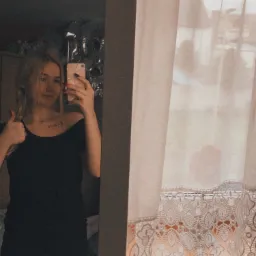 Młoda kobieta z jasnymi włosami w czarnym ubraniu robi selfie w lustrze, pokazując kciuk w górę, z widocznym fragmentem pokoju i okna z koronkową firanką.