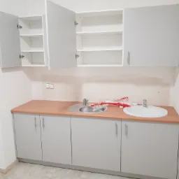 Szare szafki kuchenne z drewnianym blatem, otwarte górne szafki ukazują puste półki, widoczne ślady po remoncie w postaci folii i opakowań na blacie.