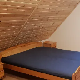 Pokój na poddaszu z drewnianą boazerią na skośnym suficie, drewnianym łóżkiem z granatową pościelą i szafkami nocnymi z lampką.
