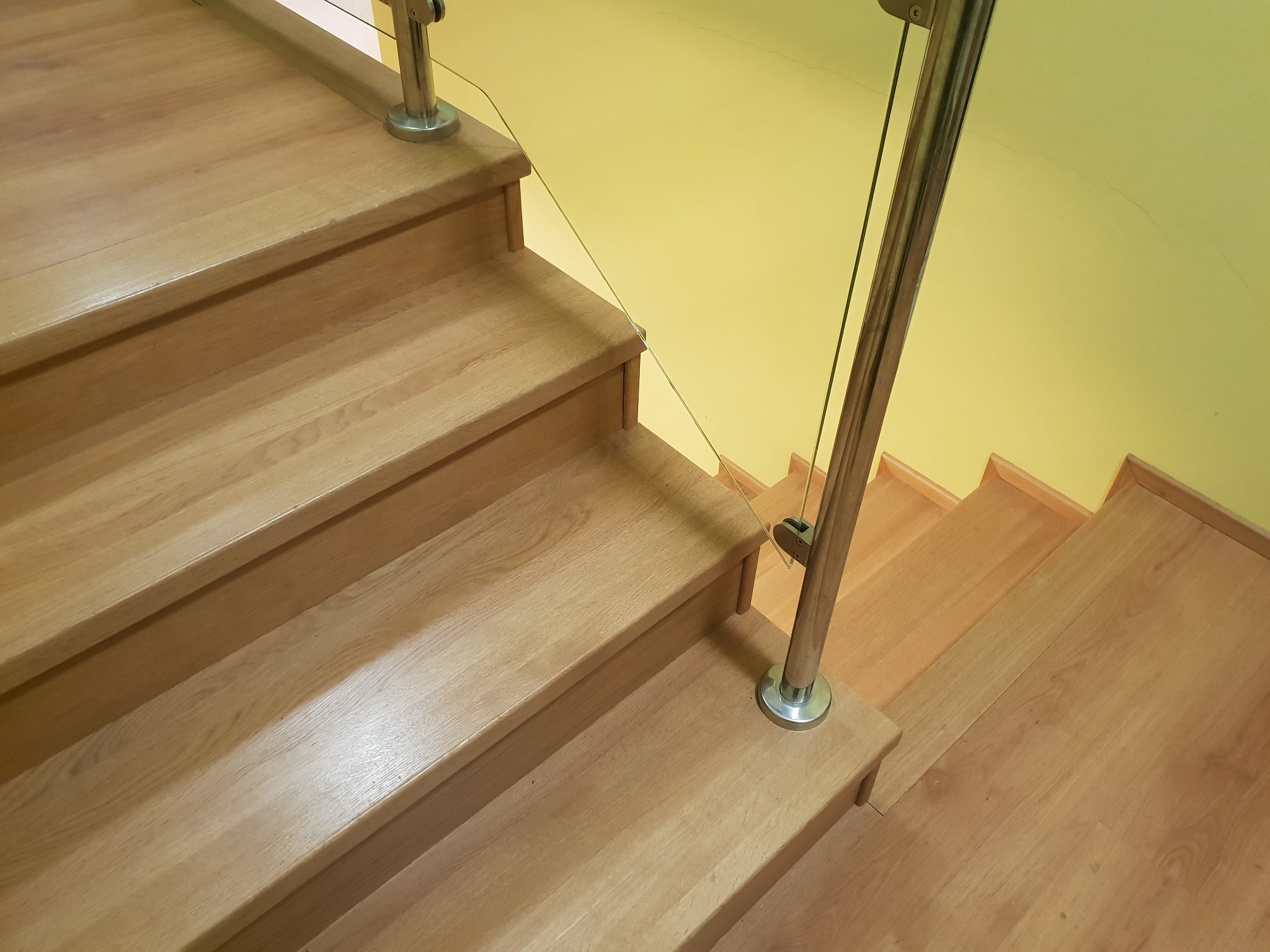 Jasne, drewniane schody z metalową balustradą i szklanym wypełnieniem, widok z góry, żółta ściana w tle.