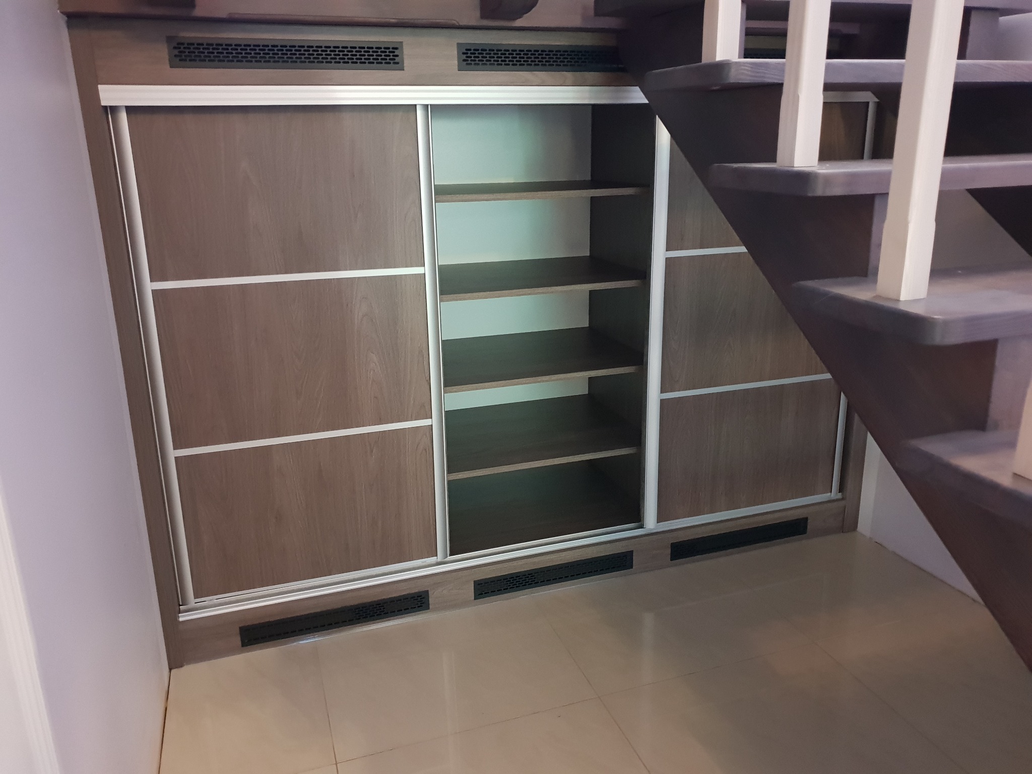 Wbudowana szafa z przesuwnymi drzwiami z okładziną imitującą drewno, z widocznymi półkami wewnątrz, częściowo zasłonięta przez drewniane schody z białymi balustradami.