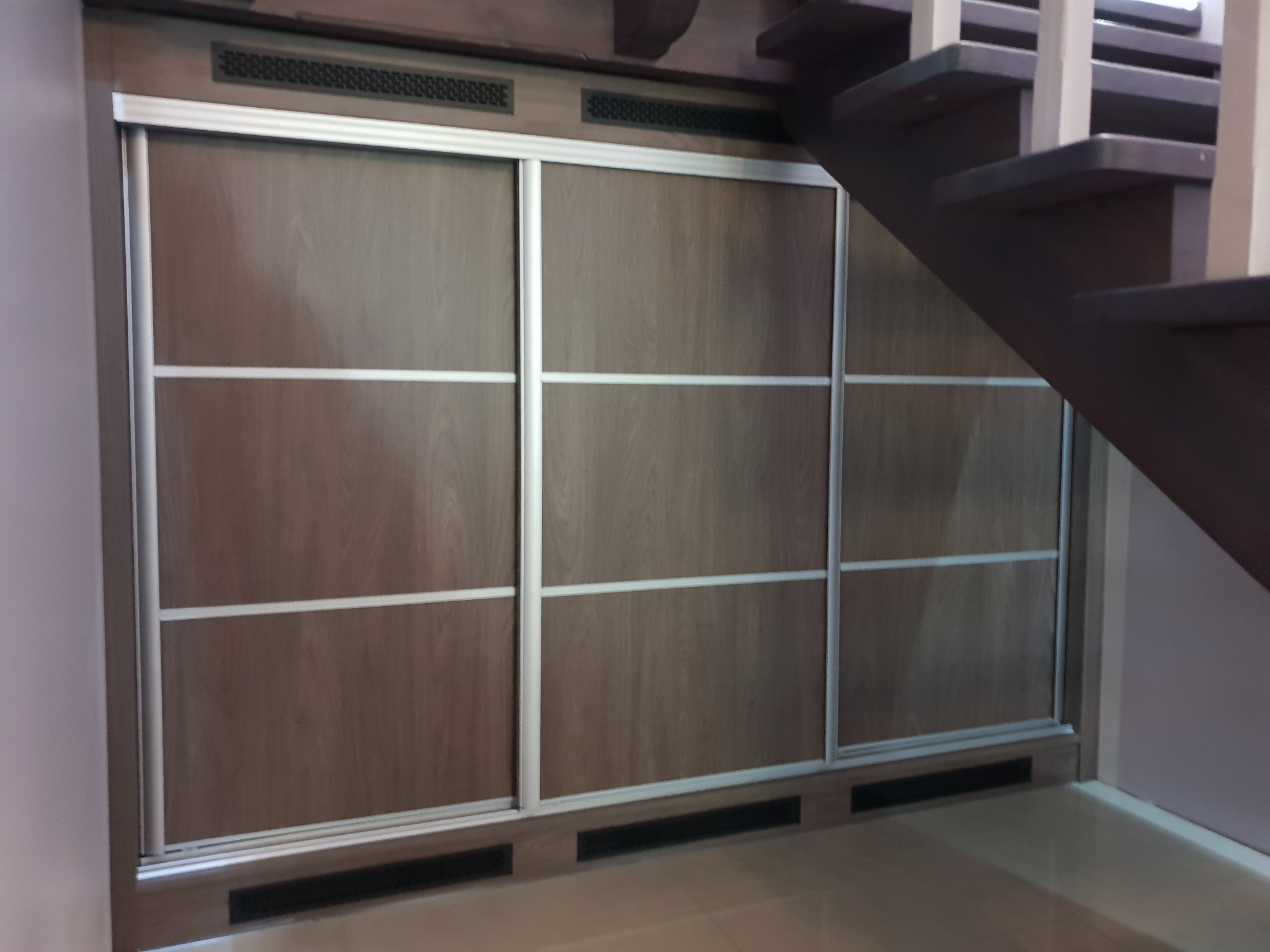 Trzydrzwiowa szafa przesuwna z drewnopodobnymi frontami i srebrnymi ramami, wkomponowana pod drewniane schody z widocznymi stopniami.