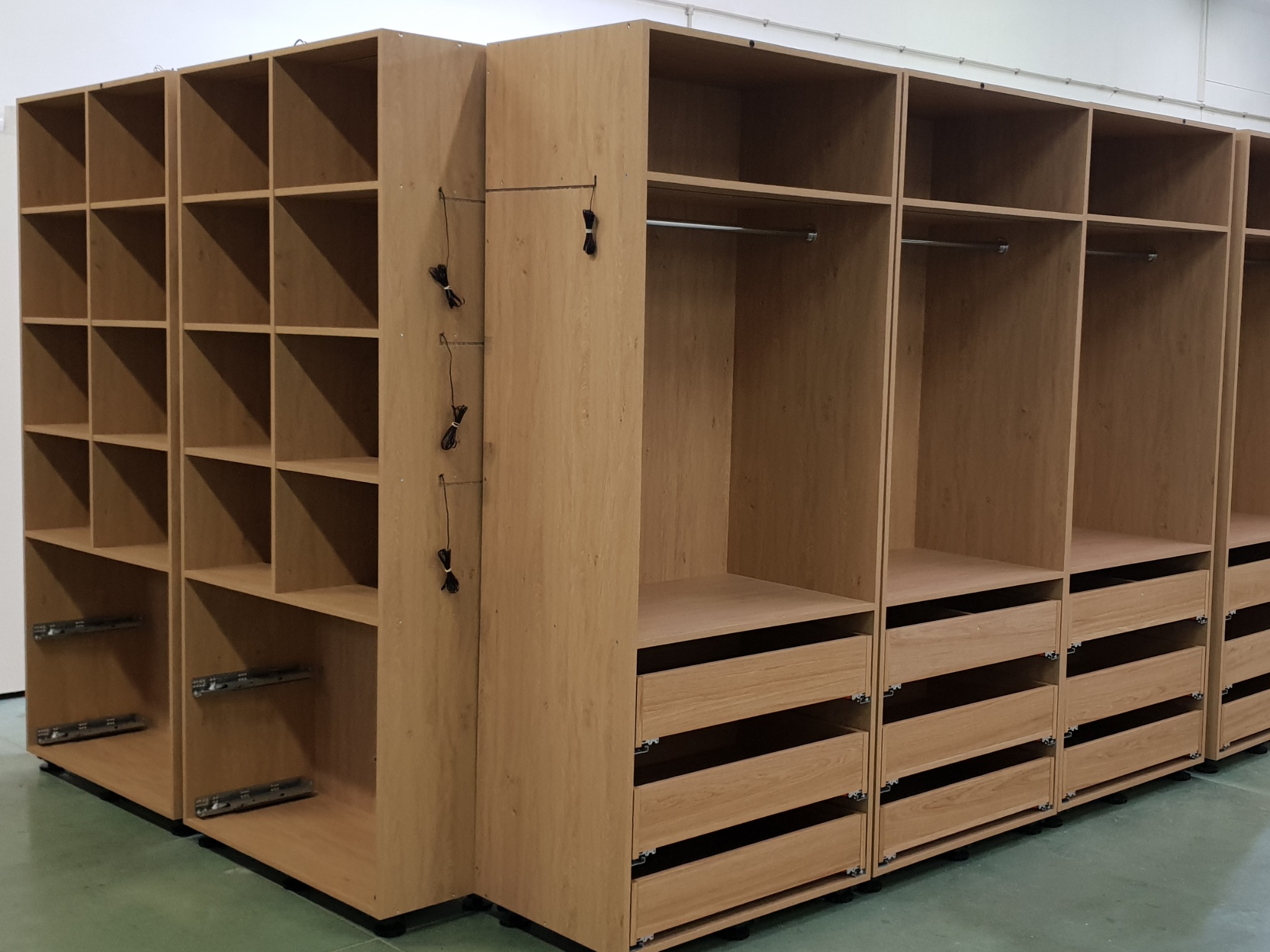 Modułowa garderoba z jasnego drewna w trakcie montażu, widoczne szuflady z systemem prowadnic, półki i drążki na ubrania, kable zwisające z boku.