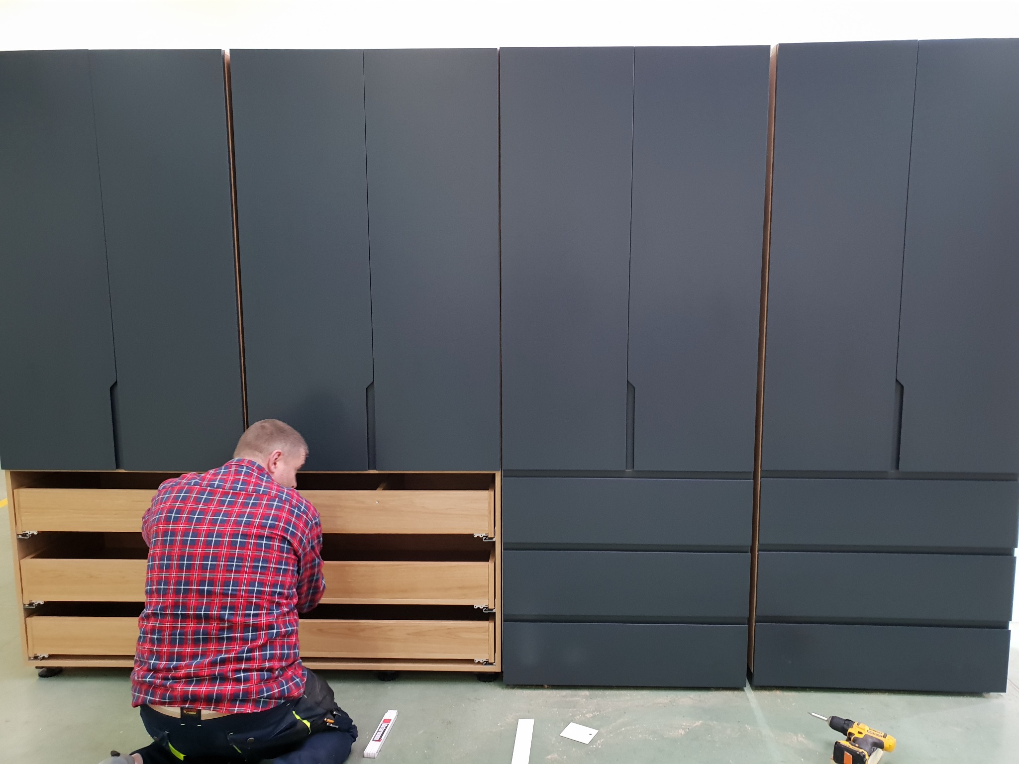 Montaż szarej szafy z drewnianymi szufladami: mężczyzna w koszuli w kratę klęczy, montując szuflady, widoczny wkrętak akumulatorowy i miarka na podłodze.